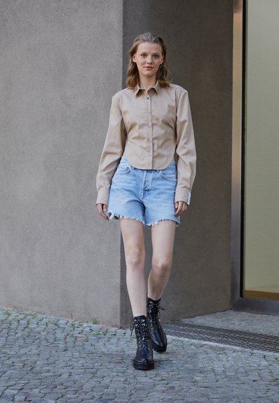 Chemise beige à boutons, shorts en denim effilochés bleu clair, et bottines noires à lacets sur une personne marchant le long d'un chemin en pierre.