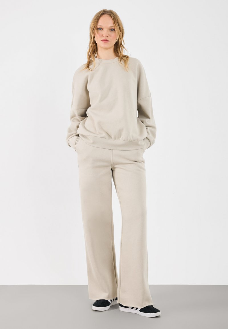 Beige sweatshirt en bijpassende wijde sweatpants gemaakt van zacht materiaal. Casual ontwerp met een ronde hals en zijzakken. Zwarte schoenen.
