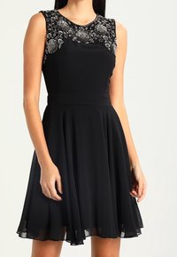 Černé šaty bez rukávů s přiléhavým živůtkem a rozšířenou sukní. Obsahují složité korálkování na výstřihu a hladkou, splývavou látku.