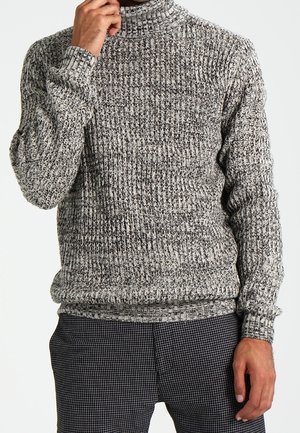 Homme portant un pull en maille gris chiné avec un pantalon à carreaux noir et blanc, la main touchant le menton, sur un fond blanc uni.