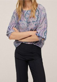 Femme les bras croisés portant un chemisier ample à manches longues à motif cachemire dans des tons violets et bleus, ainsi qu'un pantalon taille haute noir.