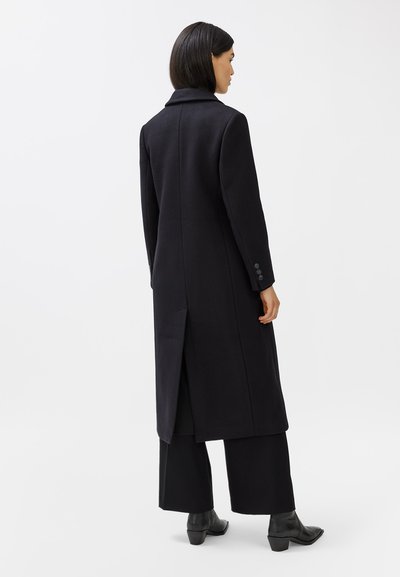 Lungo cappotto di lana nera con colletto a scollatura a V, spacco laterale e polsini abbottonati, abbinato a pantaloni neri a gamba larga e stivaletti alla caviglia.