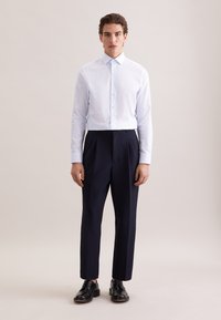 Weißes Hemd mit kleinem Punktmuster, kombiniert mit eng anliegenden dunkelblauen Hosen und schwarzen Lederschuhen. Sauberer, maßgeschneiderter Look.