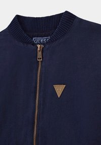 Jūrų pajėgų bombonešio striukė su raukšlėtu apykakle, užtrauktuku ir rudu odiniu trikampio logotipo ženkleliu netoli užtrauktuko. Lygus audinio paviršius.
