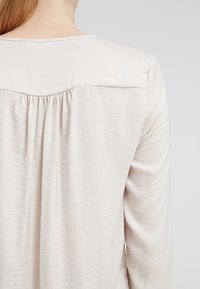 Top lungo beige chiaro a maniche lunghe con scollo rotondo, yoke posteriore arricciato e un tessuto morbido e leggermente testurizzato.