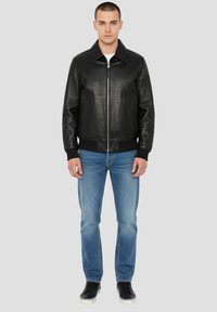 Schwarze Leder-Bomberjacke mit durchgehendem Reißverschluss, klassischem Kragen sowie gerippten Bündchen und Saum, kombiniert mit hellblauen Jeans und schwarzen Sneakers.