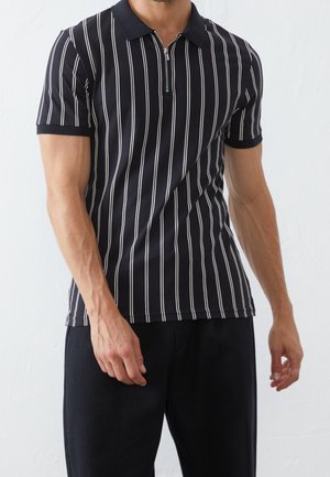 Mann trägt ein schwarzes kurzärmeliges Poloshirt mit weißen vertikalen Streifen und einem Frontreißverschluss, kombiniert mit schwarzen Hosen.