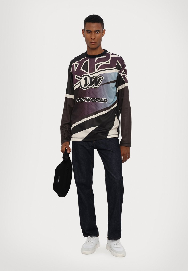 Langärmliges Grafikshirt in dunklen Tönen mit blauer Farbverlauf, das kräftige Designs und Logos zeigt. Kombiniert mit dunklen Jeans und weißen Sneakern.
