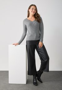 Maglione grigio a maglia con scollo a V e polsini a coste, abbinato a pantaloni wide-leg in finta pelle nera e stivaletti. Piedistallo bianco sullo sfondo.
