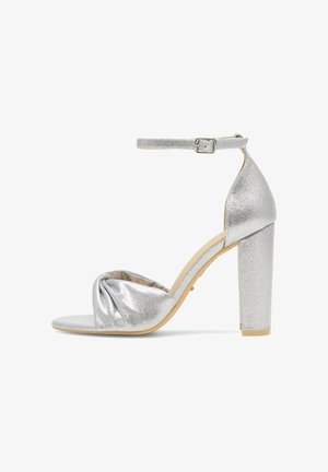 Sandalia de tacón alto con purpurina plateada, correa en el tobillo, detalle frontal anudado y tacón grueso sobre un fondo blanco.