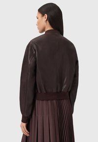 Giacca bomber in pelle marrone con polsini e orlo a coste. Texture liscia, vestibilità leggermente oversize. Cuciture dettagliate, indossata con una gonna plissettata.