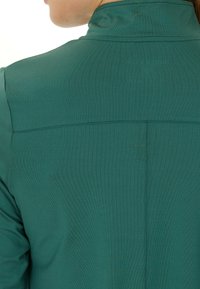 Grøn stoftop med højt krave, der har en glat overflade og diskret syninger langs ryggen. Ingen synlige lukninger.