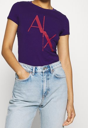Person iført en lilla Armani Exchange t-shirt med røde logobogstaver og lyseblå højtaljede jeans, den ene hånd i lommen.