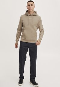 Beige hoodie med snoretræk og frontlomme, parret med mørke teksturerede bukser og sorte sneakers. Minimalistisk design med logodetalje.