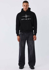 Felpa nera con logo argentato, dotata di tasca frontale. Un paio di jeans grigi scuri e larghi con una texture sbiadita. Sneakers nere.