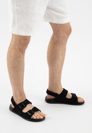 Nohy v bílých šortkách a černých sandálech s otevřenou špičkou, dvojitými přezkami a oporou paty na bílém pozadí.