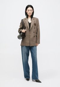 Blazer dublu-brodat în carouri maro cu detalii de blazon, asortat cu blugi închisi la culoare, pantofi negri și o geantă rotundă neagră.