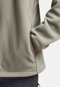 Hellgrauer Fleece-Pullover mit einer strukturierten Oberfläche, der Seitentaschen und gerippten Abschlüssen an den Bündchen und dem Saum aufweist.