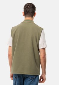 Fleece-Weste in Olivgrün, ärmelloses Design mit weicher Textur. Über einem weißen T-Shirt getragen, verfügt sie über einen geraden Saum und eine schlichte Verarbeitung.