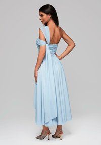 Vestito blu chiaro, off-the-shoulder, con un nodo sulla schiena e una gonna fluida. Il tessuto sembra liscio con una leggera lucentezza. Le calzature presentano un design con cinturini.