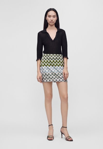 Femme debout portant un haut noir à manches longues avec une mini-jupe à motifs verts, bleus et gris, et des sandales noires à talons hauts.