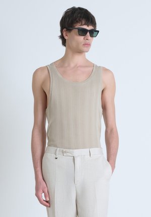Top de tirantes beige con textura acanalada y escote redondo, combinado con pantalones claros y ajustados. La modelo lleva gafas de sol negras con forma angular.