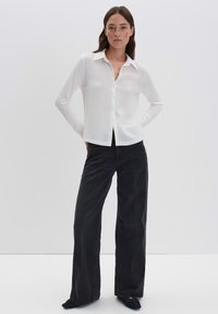 Donna in piedi con le mani dietro la schiena, indossando una camicia bianca con bottoni, jeans neri a gamba larga e scarpe nere con punta su sfondo bianco.