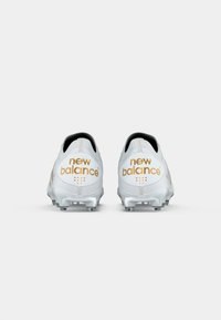Chuteiras de futebol brancas com o logo "new balance" em dourado, parte superior texturizada, detalhes em preto e cravos moldados na sola para tração.