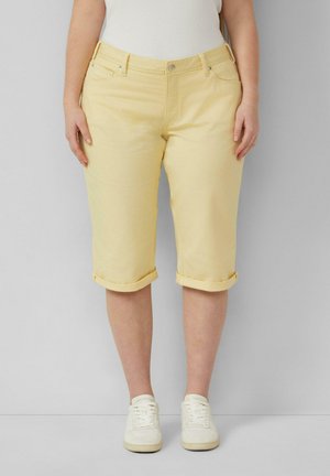 Frau trägt hellgelbe knielange Shorts mit hochgekrempeltem Saum und weißen Sneakers, steht vor einem einfarbigen grauen Hintergrund.
