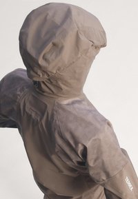 Personne portant une veste d'extérieur à capuche marron clair avec des manches texturées et le logo "TERREX" sur le bas de la manche, vue de dos sur un fond uni.