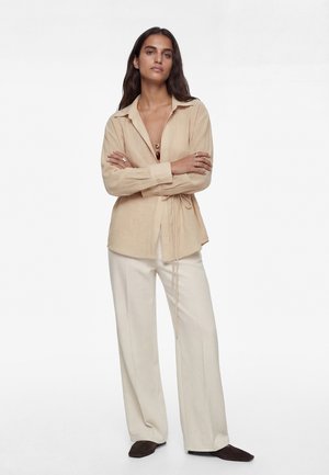 Kobieta o długich ciemnych włosach, ubrana w beżową bluzkę kopertową, białe szerokie spodnie, naszyjniki warstwowe oraz ciemne szpiczaste buty, stoi z założonymi rękami.