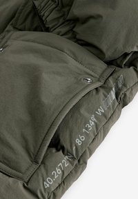 Giacca isolante verde oliva con una texture liscia, bottoni a pressione e coordinate stampate sul lato sinistro. Design leggermente trapuntato.