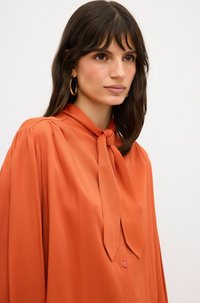 Orange Bluse mit gebundenem Kragen, weichem Stoff, langen Ärmeln, leichter Textur und Knopfverschluss, im lässigen Schnitt.