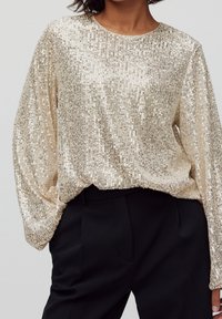 Haut à manches longues en perles dorées avec une coupe décontractée, un col rond et une surface texturée, associé à un pantalon noir ajusté.