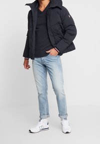 Schwarze Pufferjacke mit Kapuze, Reißverschluss vorne und weiß-roten Akzenten an den Bündchen. Getragen über einem dunklen Pullover und hellblauen Jeans, kombiniert mit weißen Sneakers.