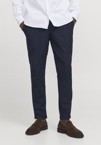 Schmal geschnittene, taillierte marineblaue Hose aus leichtem Material, mit seitlichen Taschen und umgeschlagenen Säumen. Kombiniert mit braunen Wildlederschuhen.