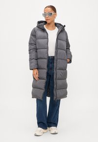Cappotto lungo grigio imbottito con cappuccio, caratterizzato da sezioni trapuntate orizzontali. Abbinato a jeans a gamba larga blu e sneakers bianche.