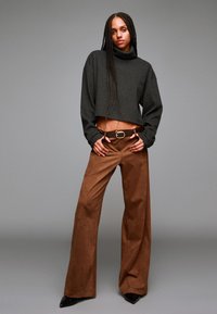 Temno siva skrajšana turtlenecka majica v kombinaciji z rjavim širokima hlačami z visokim pasom in črnimi petami s koničastim nosom. Gladek material, minimalne podrobnosti.
