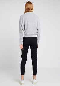Maglione grigio lavorato a maglia con una vestibilità morbida, abbinato a pantaloni neri a gamba stretta e sneakers bianche. Lo sfondo è neutro e privo di ingombri.