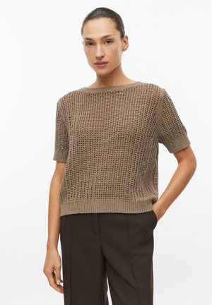 Frau mit zurückgebundenem Haar, die einen kurzärmeligen braunen Strickpullover und dunkelbraune Hose trägt, mit einer Hand in der Tasche.