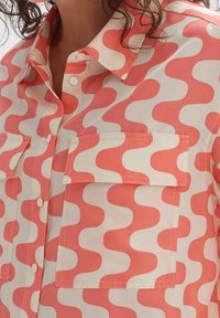 Shirt with coral and white wavy pattern, featuring front flap pockets and white buttons on a collared design.