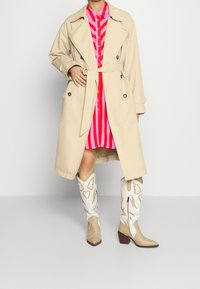 Person wearing beige trench coat over a pink and red striped dress, paired with beige and white embroidered cowboy boots.
