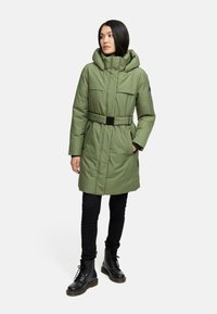 Grøn puffervinterjakke med bælte i taljen, hætte og store forlommer, iført over sorte skinny jeans og sorte snørebåndsstøvler.