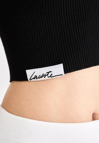 Top scurt, negru, cu dungi, având o etichetă albă cu un branding în stil scris de mână pe tiv. Partea de jos este realizată dintr-un material alb neted.