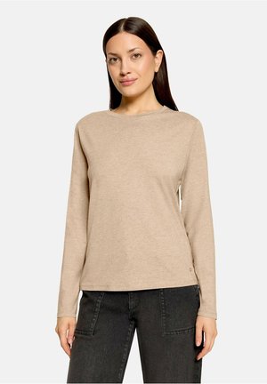 Frau mit glatten dunklen Haaren, die ein beiges Langarmshirt und schwarze Hose trägt, steht vor einem schlichten hellen Hintergrund.