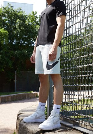 Schwarzes T-Shirt mit kurzen Ärmeln, weiße Shorts mit schwarzem Nike-Logo, weiße High-Top-Sneaker und weiße Socken mit schwarzem Logo.