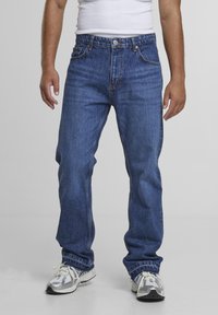 Bărbat purtând blugi albaștri largi, adidași albi și un tricou alb fără mâneci, stând în picioare pe un fundal gri simplu.