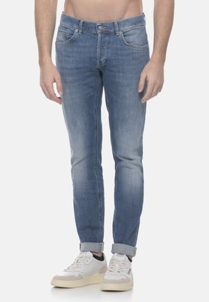 Uomo con jeans azzurri chiari risvoltati e sneakers bianche in piedi su uno sfondo semplice.