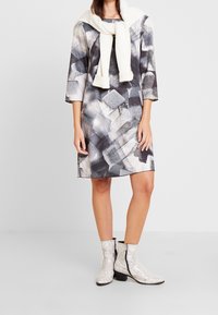 Vestido estampado en tonos de gris y crema, con mangas de tres cuartos, combinado con un suéter de punto blanco caído sobre los hombros y botas de piel de serpiente.