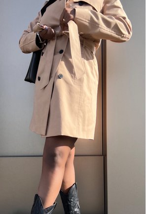 Personne portant un trench-coat beige en train de nouer la ceinture, bottes de cow-boy noires et tenant un sac à main noir devant un mur neutre.